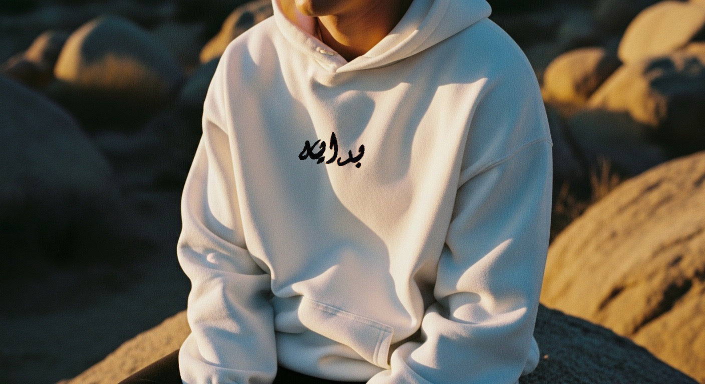 Person wearing a white hoodie with Arabic text on a beach at sunset